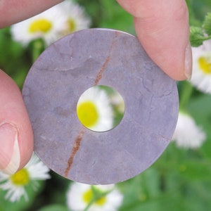 Peut inclure: Un pendentif en forme de beignet en pierre violette et brune avec un trou au centre. Le pendentif est tenu par une main sur un fond de fleurs blanches.