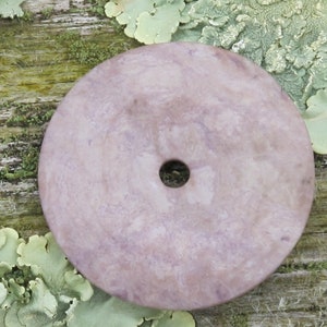 May include: A round, flat, purple and white stone disc with a hole in the center. The disc is lying on a mossy surface.