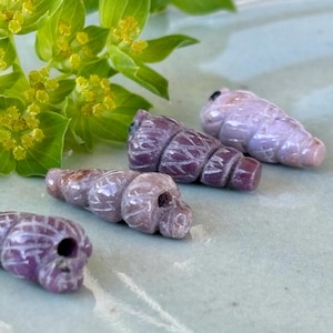 May include: Four carved purple gemstone beads in the shape of seashells. The beads are a mix of light and dark purple hues, with detailed carved lines. The beads are displayed on a light green surface, with green foliage in the background.