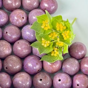 May include: Close-up of a collection of round, mauve-colored beads with drilled holes, arranged closely together. A sprig of green leaves with small yellow flowers rests among the beads, adding a natural contrast to the smooth, polished surfaces.