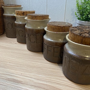 May include: A set of five cylindrical ceramic storage canisters with cork lids. Each canister features a two-tone glaze with a light cream top and a dark brown base. Embossed lettering on the front of the canisters includes the words RICE, COFFEE, and TEA.