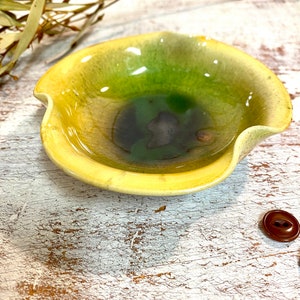 May include: A small, yellow ceramic bowl with a green and blue glaze. The bowl has a wavy edge and is sitting on a white wooden surface.