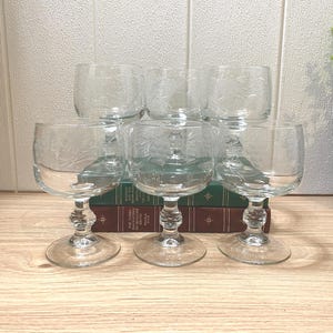 May include: A set of six clear glass goblets with short stems and round bases. Each goblet features a delicate etched design of pine cones and needles on the bowl. The glasses are arranged in two rows, with three glasses in the front and three in the back.