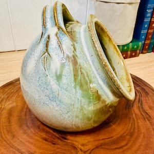 May include: A round ceramic salt pig with a wide, angled opening and a single handle. The vessel features a mottled glaze in shades of light blue, sage green, and cream, with brown accents along the rim and handle, showcasing a smooth, glossy finish.