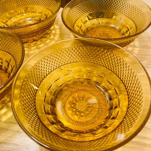May include: A collection of amber-coloured glass bowls featuring a diamond pattern. The bowls are round with a decorative centre design. They are arranged on a wooden surface, with books visible in the background.