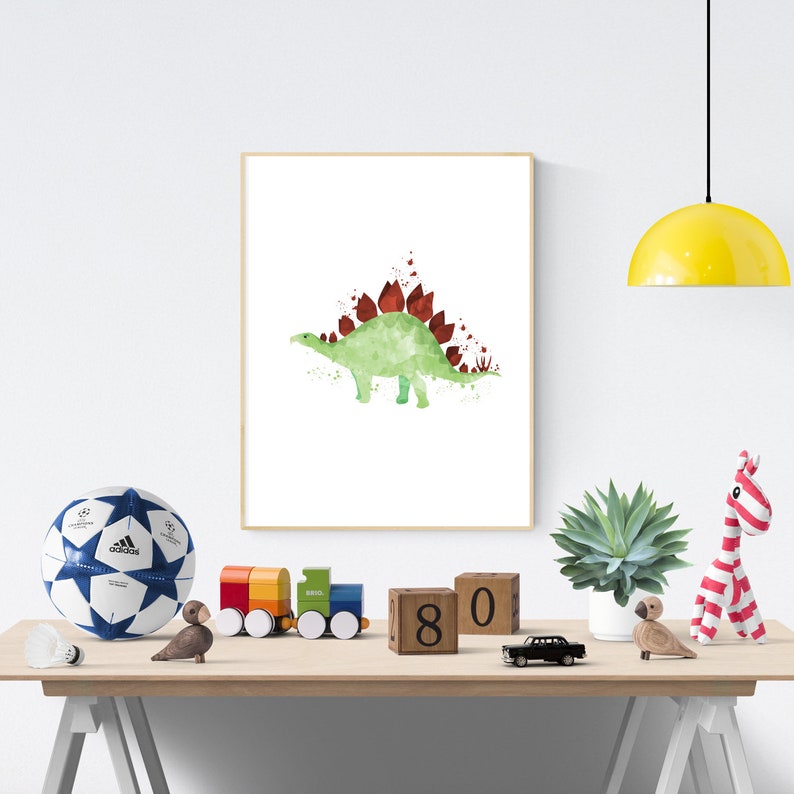 May include: A watercolor print of a green dinosaur with red spines. The dinosaur is standing on a white background. The print is framed and hanging on a white wall.