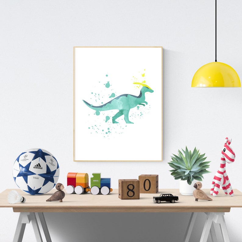 May include: A watercolor print of a green dinosaur with a yellow crown, surrounded by blue and green paint splatters. The print is framed and hanging on a white wall.