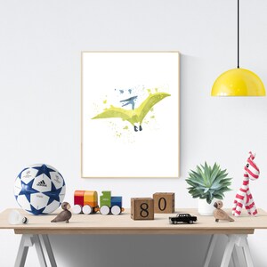 May include: A watercolor print of a green pterodactyl flying with a blue sky and white clouds in the background. The print is framed and hanging on a white wall.