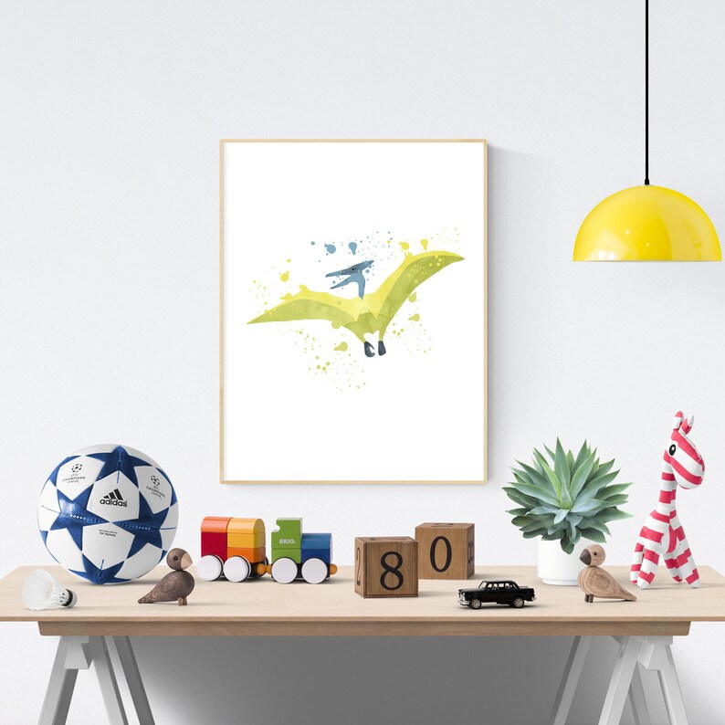 May include: A watercolor print of a green pterodactyl flying with a blue sky and white clouds in the background. The print is framed and hanging on a white wall.