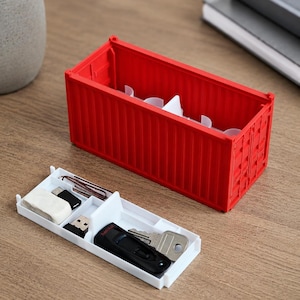 May include: A red container-shaped desk organizer with a white tray. The tray holds a black USB drive, keys, a USB-C adapter, an eraser, and paperclips. The organizer sits on a wooden surface.