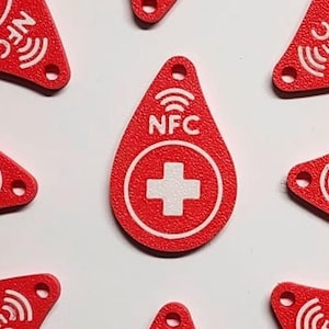 May include: Red NFC tags with a white cross symbol and the letters "NFC". The tags are teardrop-shaped with a hole at the top. The tags are scattered on a white surface.