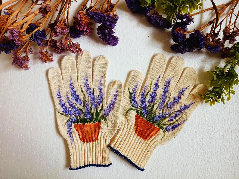 May include: A pair of beige gardening gloves with a painted design of lavender in orange pots. The gloves have a ribbed cuff with a navy blue trim. Dried purple flowers and green foliage are in the background.