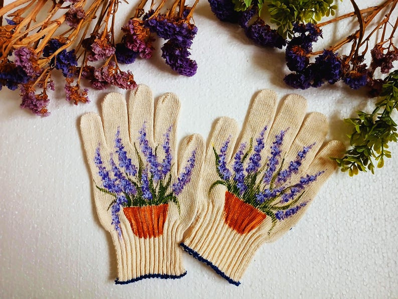 May include: A pair of beige gardening gloves with a painted design of lavender in a terracotta pot. The gloves have a ribbed cuff with a navy blue trim. Dried flowers and greenery are in the background.