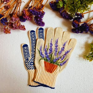 May include: Cream-colored gardening gloves with a lavender plant design. The gloves have blue grip patterns on the palms and fingertips. The lavender is in a terracotta pot. Dried flowers and greenery are in the background.