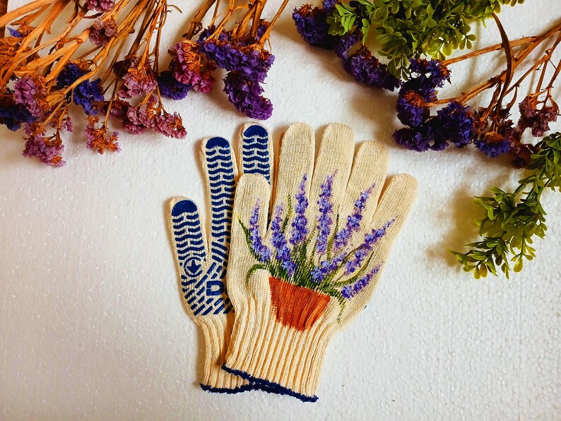 May include: Cream-colored gardening gloves with a lavender plant design. The gloves have blue grip patterns on the palms and fingertips. The lavender is in a terracotta pot. Dried flowers and greenery are in the background.
