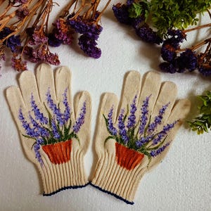 May include: A pair of beige knitted gloves with a detailed painting of lavender plants in orange pots. The lavender has purple flowers and green leaves. The gloves have a ribbed cuff with a dark blue trim.