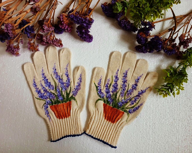 May include: A pair of beige knitted gloves with a detailed painting of lavender plants in orange pots. The lavender has purple flowers and green leaves. The gloves have a ribbed cuff with a dark blue trim.