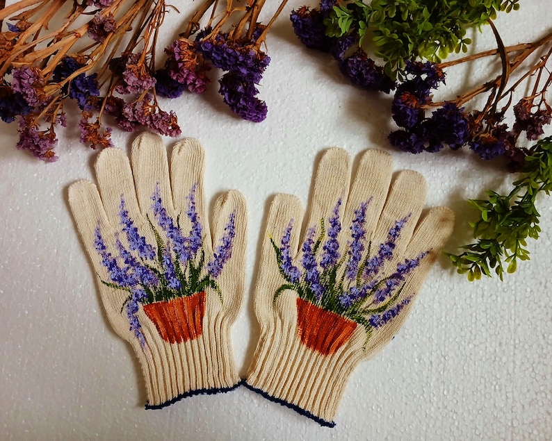 May include: A pair of beige knitted gloves with a detailed painting of lavender plants in orange pots. The lavender has purple flowers and green leaves. The gloves have a ribbed cuff with a dark blue trim.