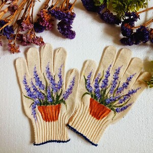 May include: A pair of beige gardening gloves with a painted design of lavender in a terracotta pot. The gloves have a ribbed cuff with a navy blue trim. Dried flowers and greenery are in the background.