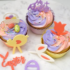 Puede incluir: Tres cupcakes de Pascua decorados con glaseado morado y rosa, chispas y varios adornos con temática de Pascua, incluyendo un pájaro, un conejito, un tulipán y una cesta. Los cupcakes están en forros de papel blanco y tienen un esquema de color amarillo y morado.
