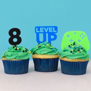 May include: Three green cupcakes with blue frosting and colorful sprinkles. The cupcakes are topped with a blue "Level Up" sign, a black number "8" and a green video game controller.