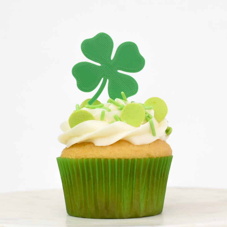 May include: A green cupcake with white frosting and green sprinkles topped with a green four-leaf clover