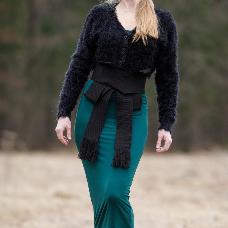 May include: A black fuzzy cropped cardigan with a black wide belt tied in a bow at the front. The belt has long fringed ends. The cardigan is worn over a teal green long-sleeved dress.