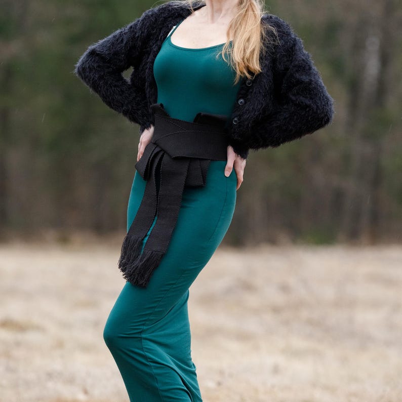 May include: A woman wearing a teal green dress with a black wide belt tied in a bow. She is wearing a black fuzzy cardigan.