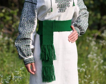 May include: A white linen dress with a green embroidered belt and a long green fringe. The dress has a traditional Ukrainian design.