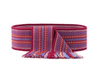 May include: A woven belt in shades of burgundy, orange, and blue. The belt features a repeating pattern of horizontal stripes and a fringed end. The belt is designed to be worn around the waist.