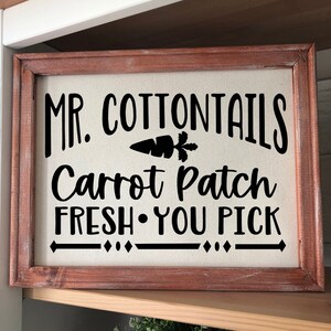 May include: A rustic wooden framed sign with a white canvas background. The sign reads "Mr. Cottontails Carrot Patch Fresh You Pick" with a black silhouette of a carrot with green leaves.
