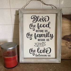 Pode incluir: Um sinal de madeira branco emoldurado com caligrafia preta que diz "Bless the food before us the family beside us and the love between us".
