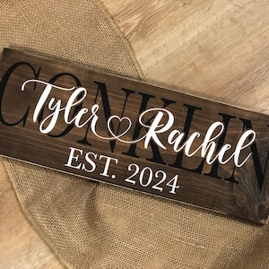 May include: A rectangular dark brown wooden sign featuring the black text CONKLIN in the background, with the white script names Tyler and Rachel separated by a heart symbol in the center, and the white text EST. 2024 printed at the bottom.