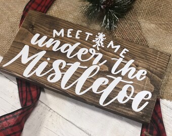 May include: A rustic wooden sign with the text "Meet me under the mistletoe" in white lettering. The sign has a distressed wood finish and a natural wood grain.
