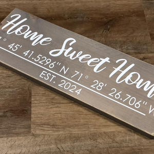 Könnte beinhalten: Ein rustikales Holzschild mit dem Text "Home Sweet Home" in weißen Buchstaben. Das Schild enthält auch die Koordinaten "41°45' 41.5296" N 71°28' 26.706" W" und das Jahr "EST. 2024".