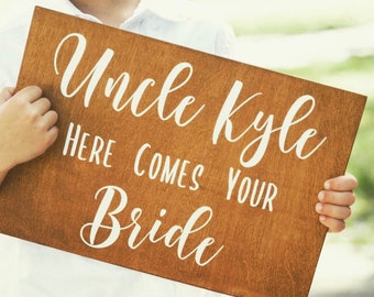 May include: A brown wooden sign with white text that reads "Uncle Kyle Here Comes Your Bride".