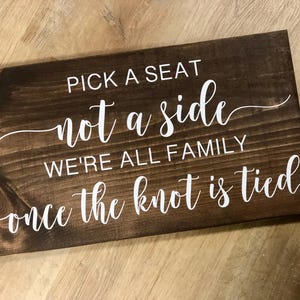 May include: A rectangular wooden sign with a dark brown stain. White cursive text reads "PICK A SEAT not a side WE'RE ALL FAMILY once the knot is tied."