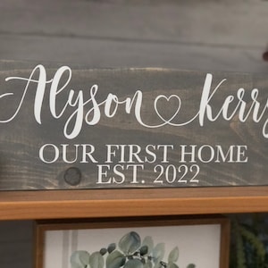 May include: A gray wooden sign with white lettering that reads "Alyson & Kerry Our First Home Est. 2022".