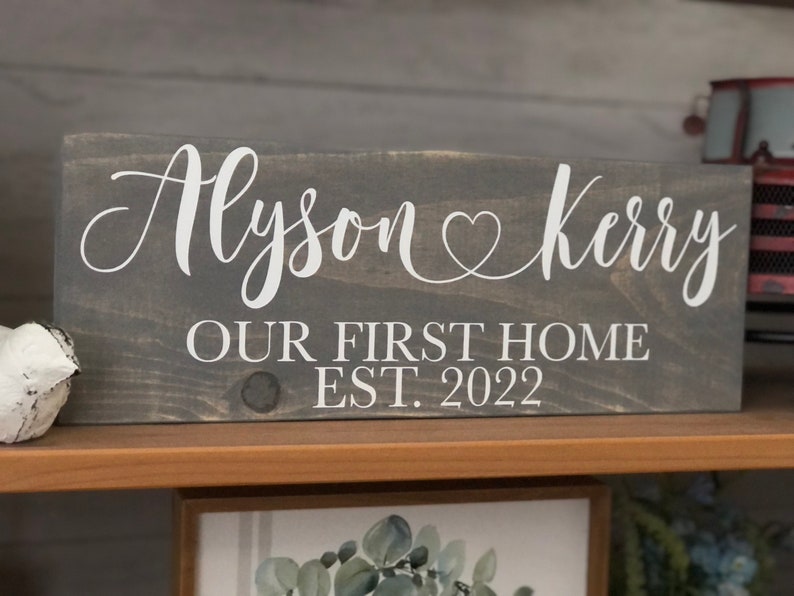 May include: A gray wooden sign with white lettering that reads "Alyson & Kerry Our First Home Est. 2022".