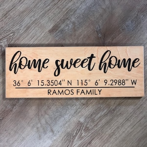 May include: A rectangular wooden sign with the words "home sweet home" in black script. Below the text are coordinates and the name "RAMOS FAMILY" in black. The sign is on a light wood surface.