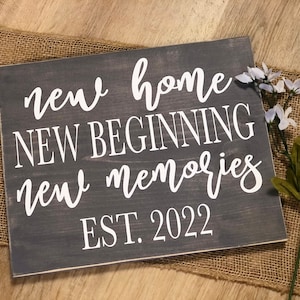 May include: A gray wooden sign with white text that reads "new home new beginning new memories EST. 2022".