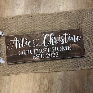 May include: A brown wooden sign with white text that reads "Artie & Christine Our First Home Est. 2022".