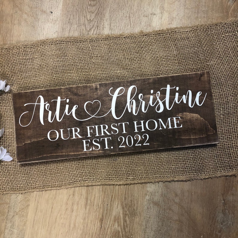 May include: A brown wooden sign with white text that reads "Artie & Christine Our First Home Est. 2022".