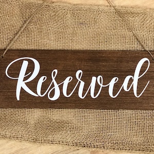 May include: A dark brown wooden sign with the word "Reserved" in white script. The sign is suspended by twine. The background is a coarse burlap material, creating a rustic aesthetic.