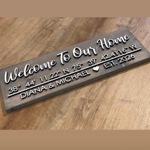 May include: A rectangular grey wooden sign featuring raised white lettering that reads Welcome To Our Home, followed by the coordinates 38 44 11.22 N 75 39 42.4116 W, a heart symbol, the text EST. 2026, and the names DIANA & MICHAEL.