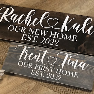 May include: Two wooden signs with white text. The top sign is dark brown and reads "Rachel & Kale Our New Home Est. 2022". The bottom sign is gray and reads "Trent & Tina Our First Home Est. 2022".