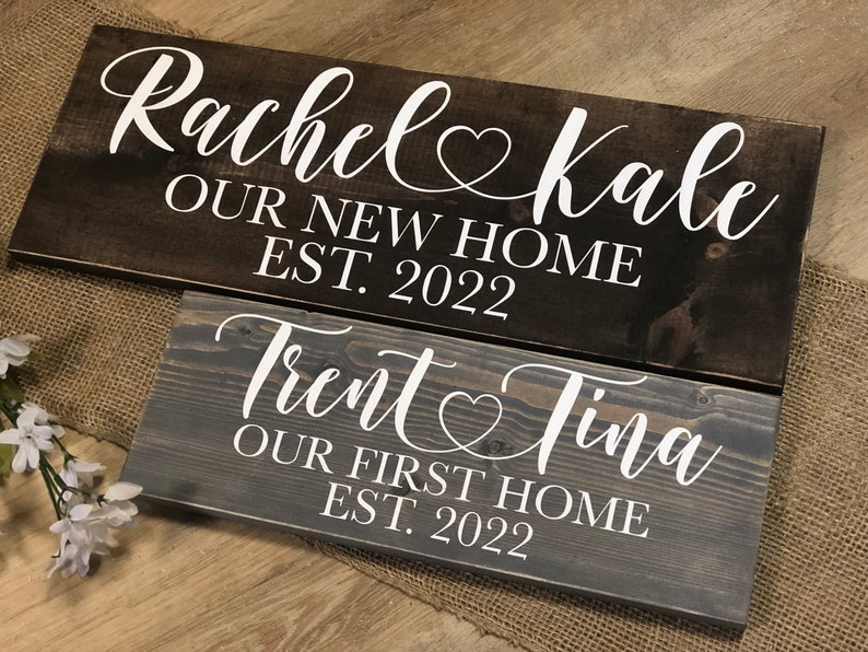 May include: Two wooden signs with white text. The top sign is dark brown and reads "Rachel & Kale Our New Home Est. 2022". The bottom sign is gray and reads "Trent & Tina Our First Home Est. 2022".