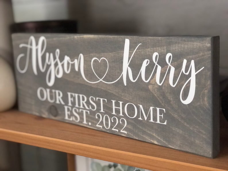 May include: A gray wooden sign with white lettering that reads "Alyson & Kerry Our First Home Est. 2022". The sign has a heart symbol between the names.