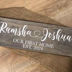 May include: A rustic wooden sign with the text "Ramsha & Joshua Our First Home Est. 2024" in white lettering. The sign features a heart symbol between the names.
