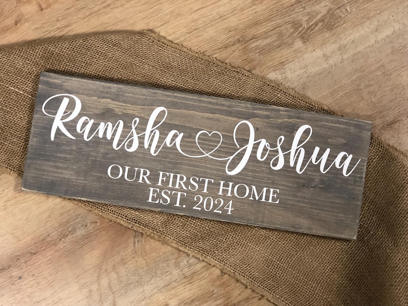 May include: A rustic wooden sign with the text "Ramsha & Joshua Our First Home Est. 2024" in white lettering. The sign features a heart symbol between the names.